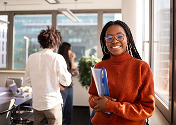 femme brune avec des tresses, porte un pull rouge-orange, des lunettes, des boucles d'oreilles, tient dans ses mains une pochette bleue (fond : salle de réunion)