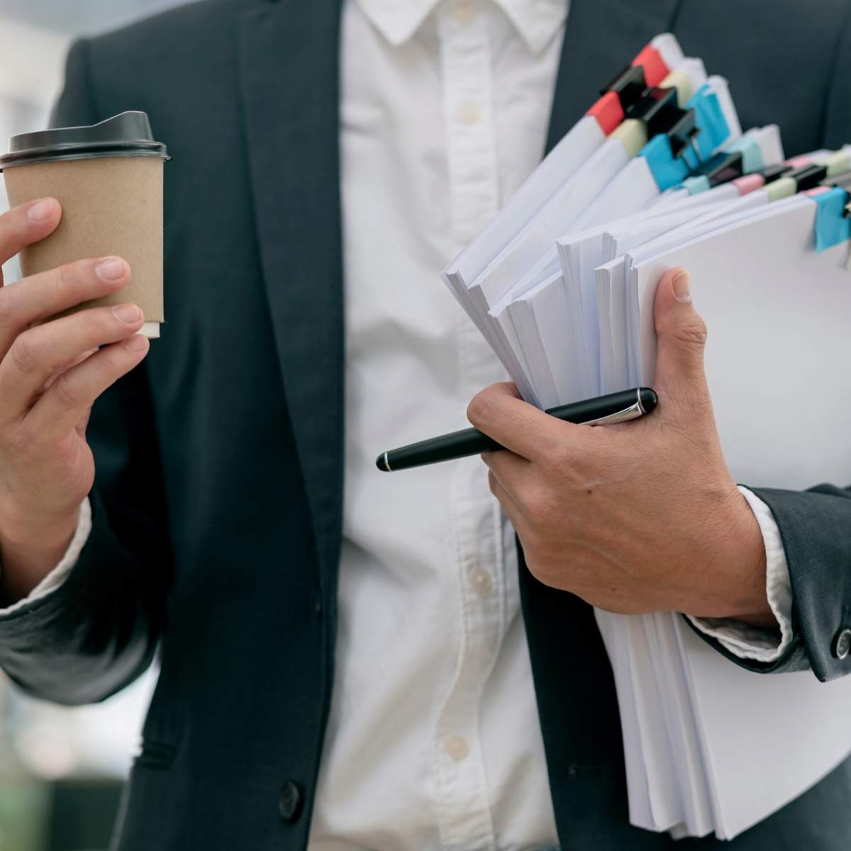 Une personne, en tailleur et chemise, porte des dossiers et un stylo de la main gauche puis un gobelet de la main droite