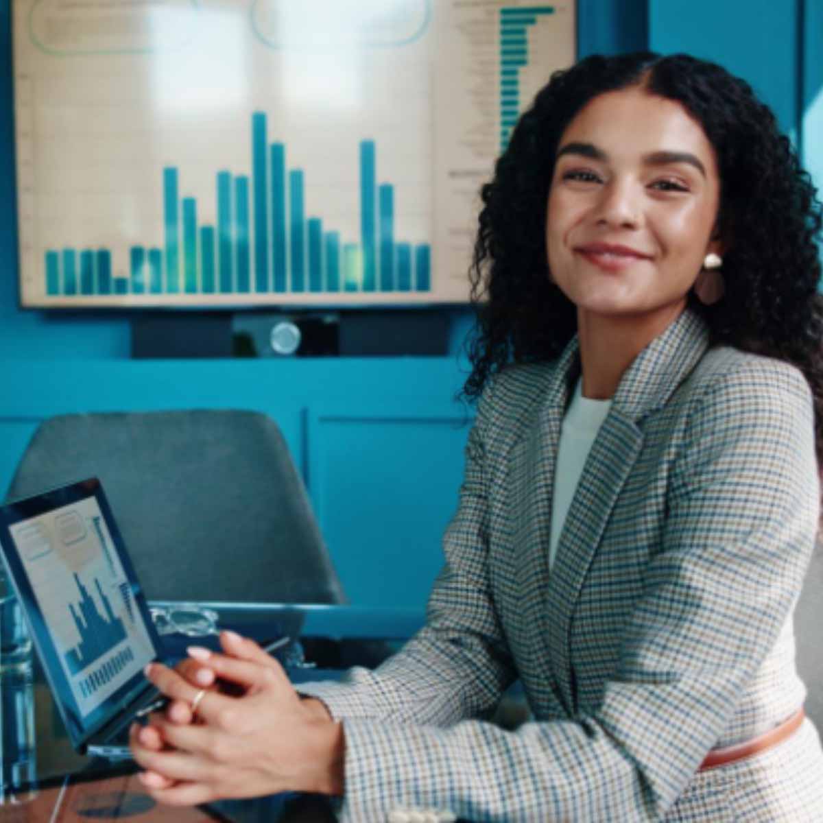 Une femme souriante regardant face caméra assise à un bureau avec dessus un ordinateur et sur le mur du fond un histogramme projeté