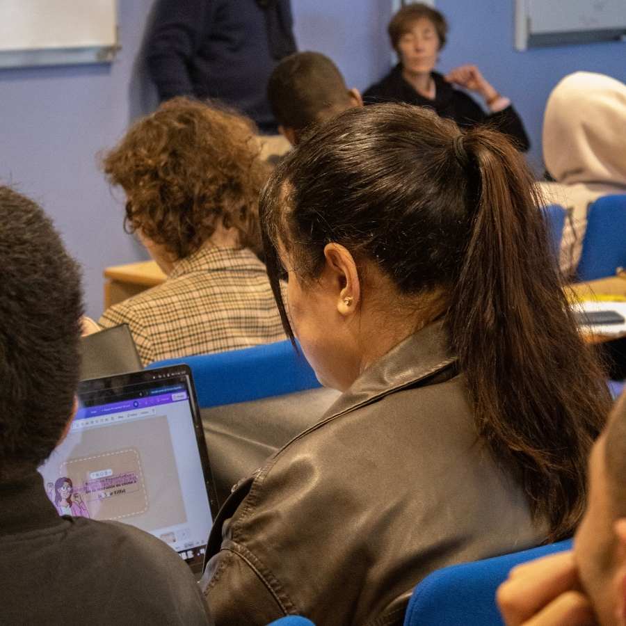 Femme brune avec les cheveux attachés assise dans une salle de classe regarde son ordinateur